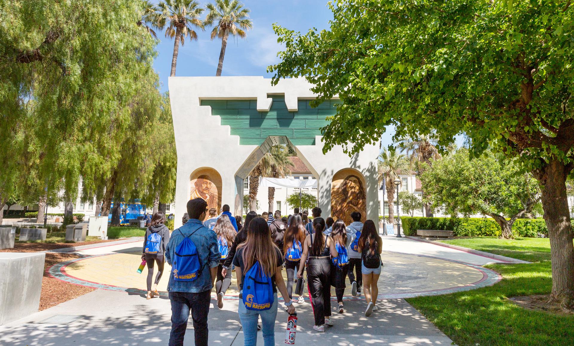 Students wearing 利记 gear are walking through the Cesar Chavez arch that is surrounded by bright green trees.